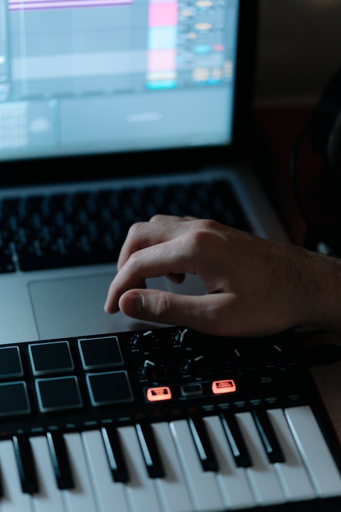 Offerings Close-up of hand adjusting synthesizer controls with laptop screen displaying audio software.